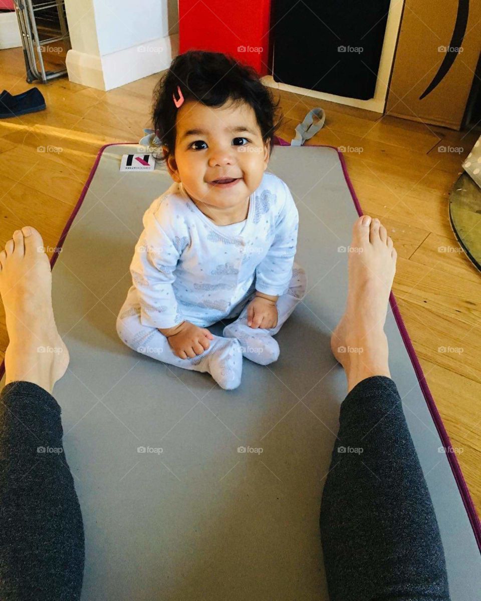 Baby in onsie, sitting up on a yoga mat between her mums feet looking directly into the camera.