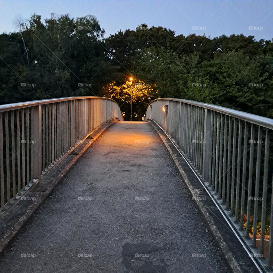 evening light on a foot bridge over the road. artificial light one end, fences, bushes