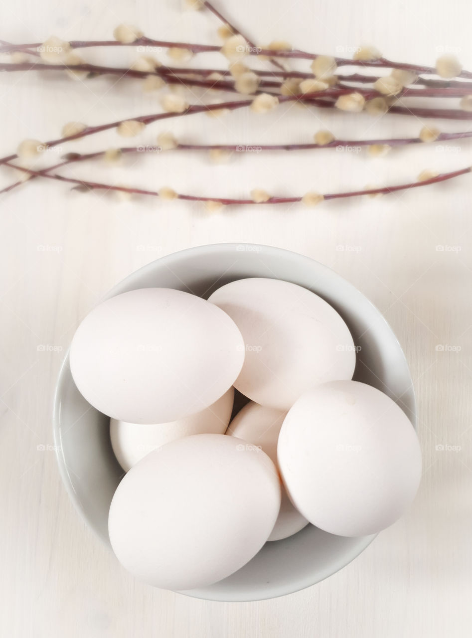 Still life with white whole raw eggs in a bowl on a white wooden table top with twigs. Preparing for the Easter holidays. Food photo, vertical orientation