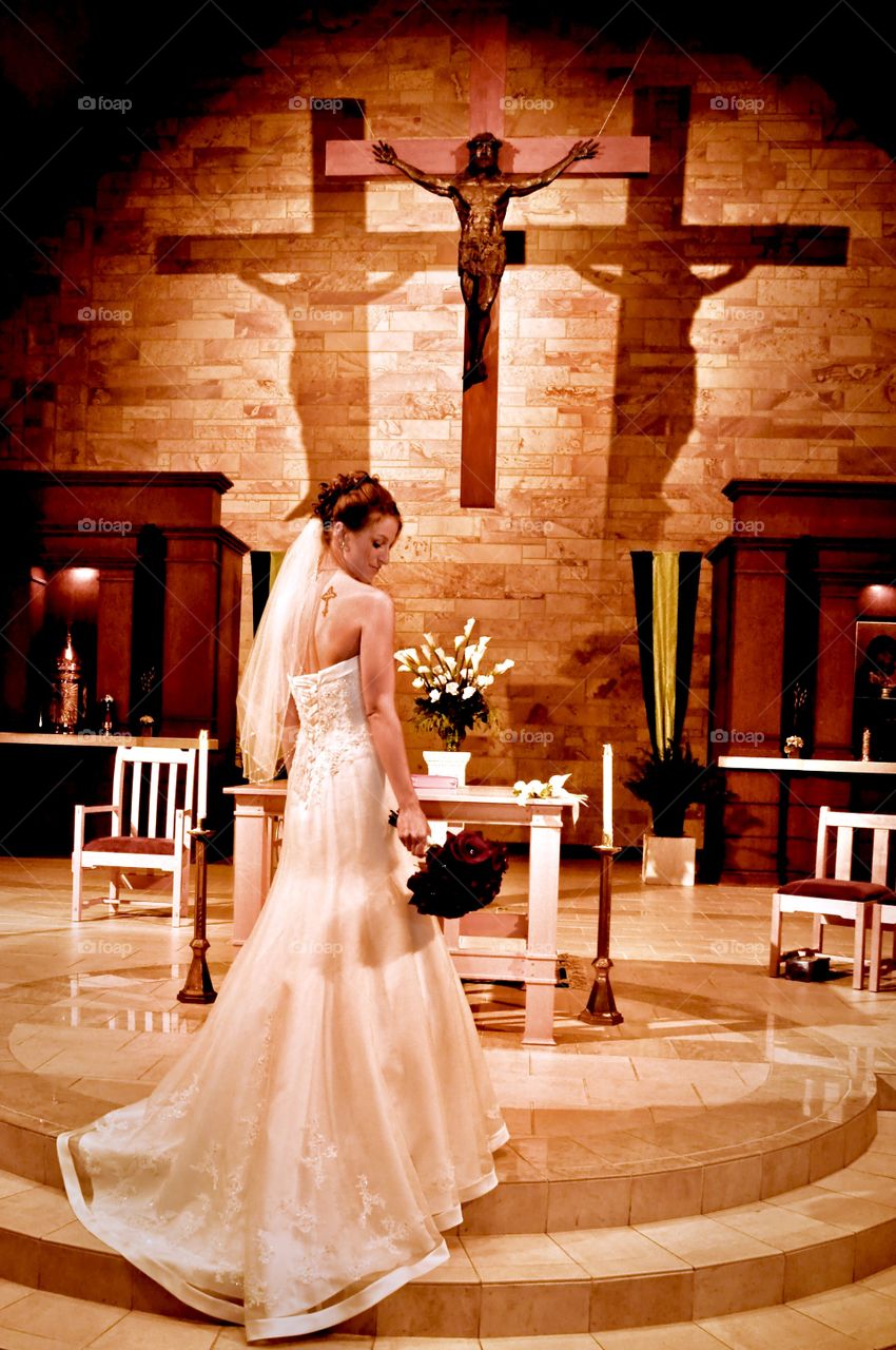 Breathtaking image of a beautiful bride captured at the alter moments before her wedding in a Catholic Church with unique lighting creating shadows behind a hanging crucifix giving an illusion of the trinity being present at mass