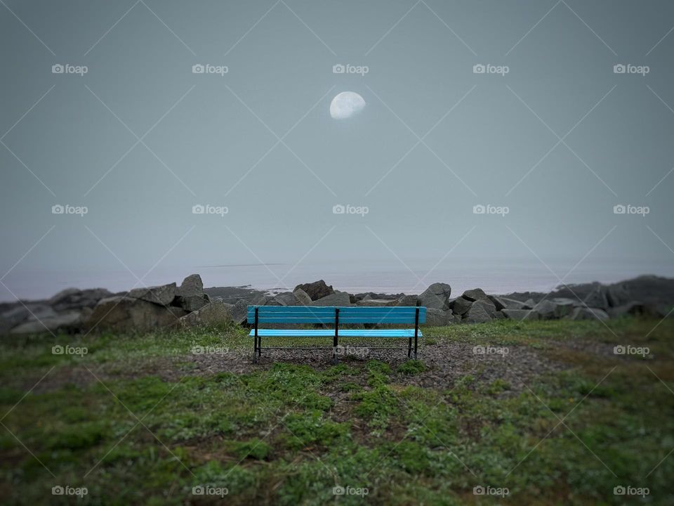 An empty blue bench overlooking water on a foggy day with a waxing moon overhead 