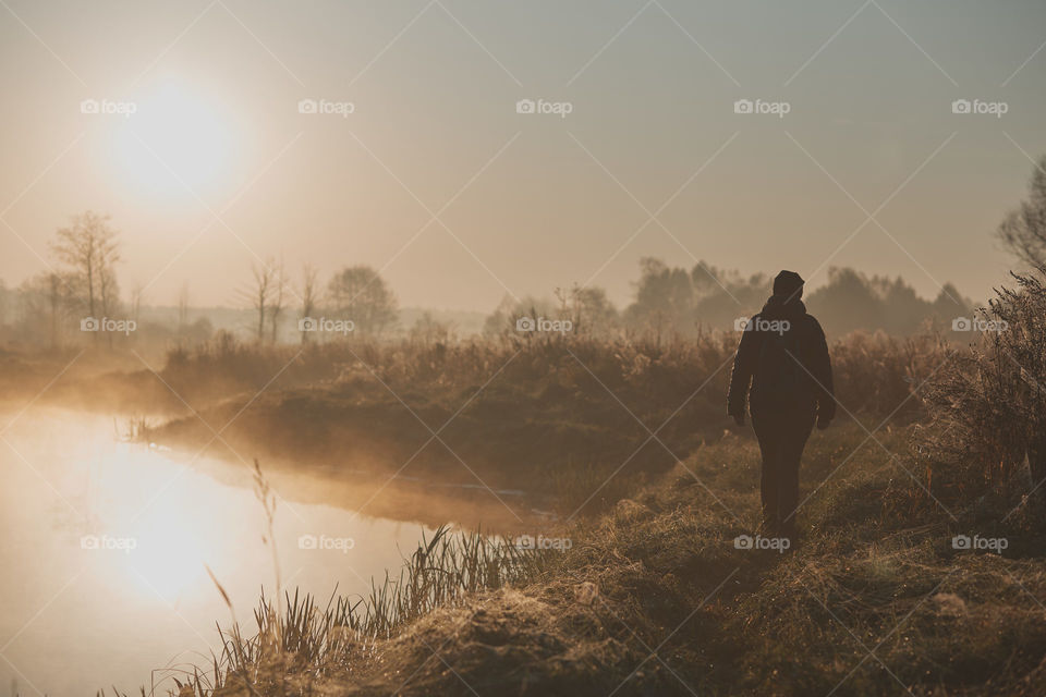 Woman walking through a meadow by a pond in the foggy morning. Sun rising above field and pond flooded with fog in the morning. Real people, authentic situations