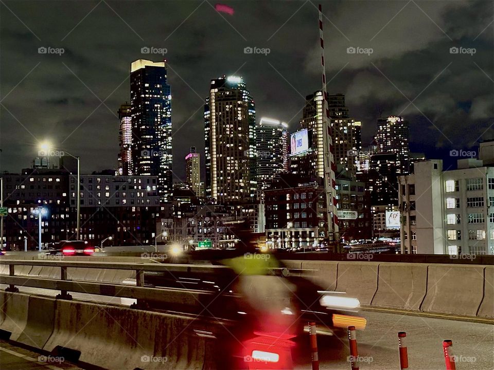 This is the bicycle lane and the car lanes of the “Pulaski Bridge” that connects “Greenpoint”, Bklyn and LIC, Queens. The dynamic of high speed is enhanced further by the trailing rear lights of a bicyclist zooming past. 2023. Hypnotic Productions
