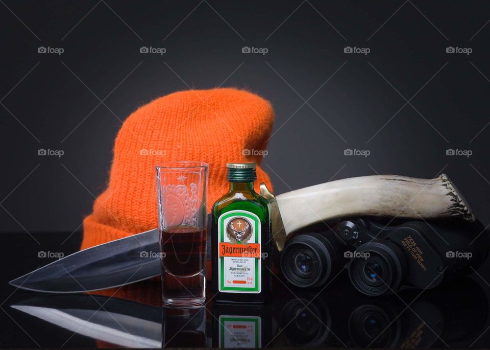 single shot bottle of liquor with half in a shot glass placed in front of an orange knit hat, hunting knife, and binoculars