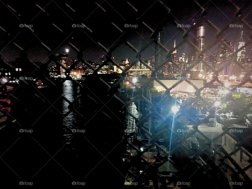 The illuminated cityscape of Long Island City, Queens, New York and of the Manhattan skyline with the Empire State Building as well as the ocean is seen through a fence standing on top of the Pulaski Bridge. Photo from 2020. Hypnotic Productions