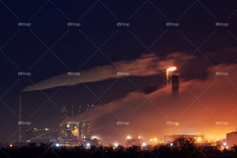 Night time over an industrial oil refinery with tall chimneys blowing smoke and steam over the image.