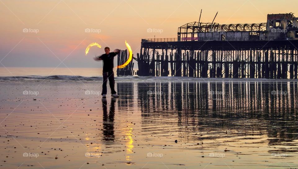 Man spinning fire poi on a wet beach, with an old pier in the background, around sunset
