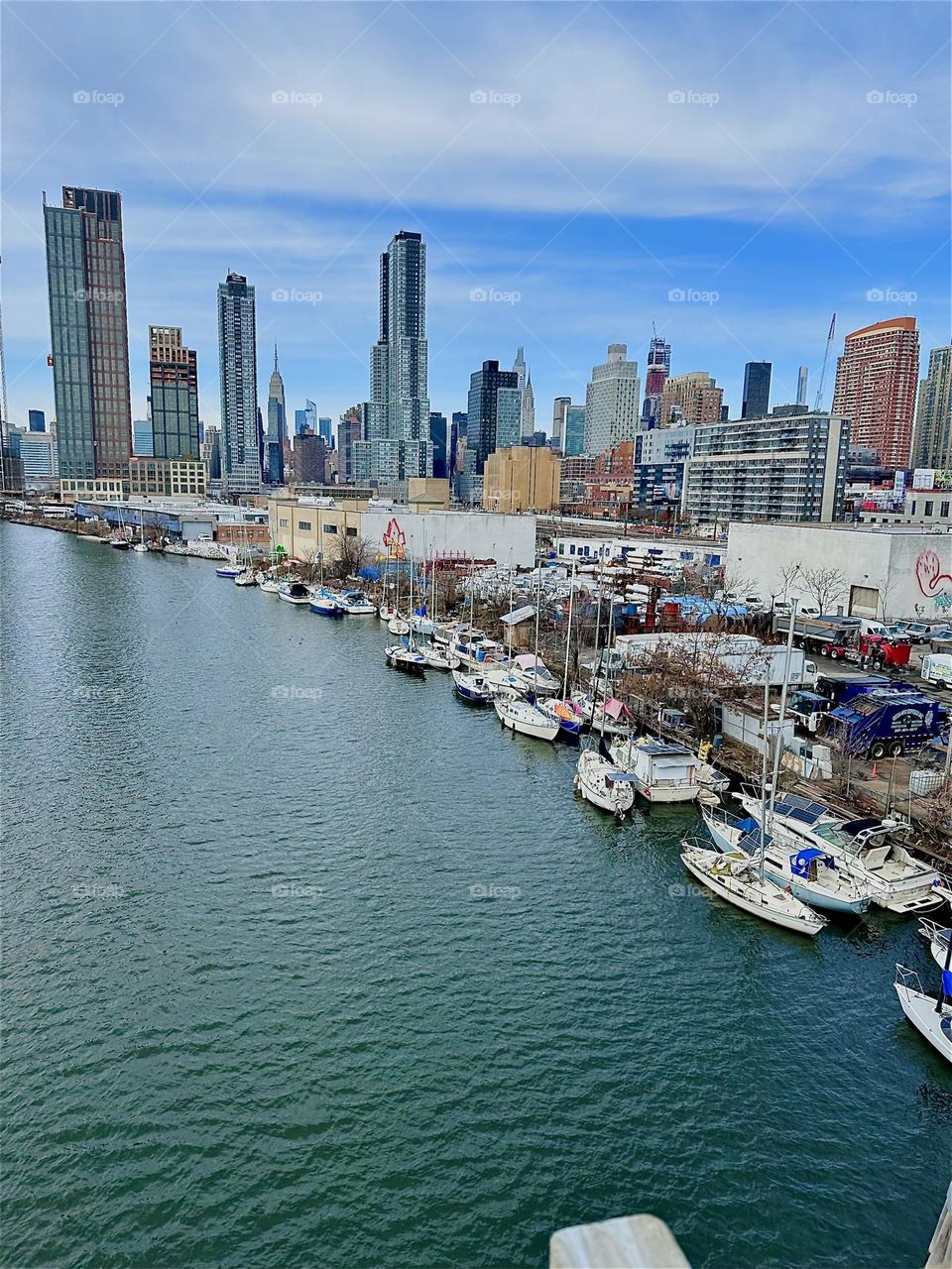 This is „Newtown Creek“ with its many boats seen from the „Pulaski Bridge“ that connects „Greenpoint“, Brooklyn to LIC, Queens. In the distance across the waters of the „East River“ we see „Manhattan“. 2024. Hypnotic Productions