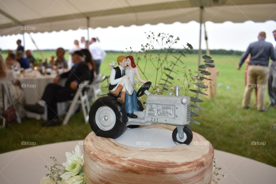 The top tier of a rustic wedding cake shows of a quirky country cake topper with cowboy boots bride and loving groom. Behind the cake topper eucalyptus and flowers hold the foreground with Wedding tent and guests in the rear. 