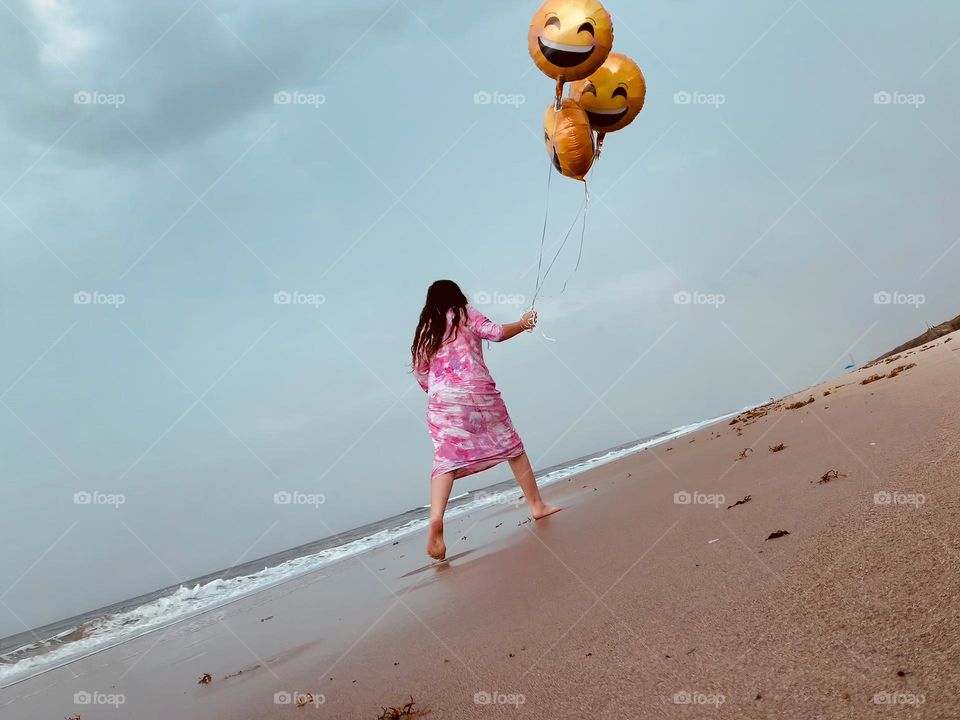 Little Girl Walking On The Sand And Having Fun With Helium Emoji’s Balloons On The Beach By The Ocean.