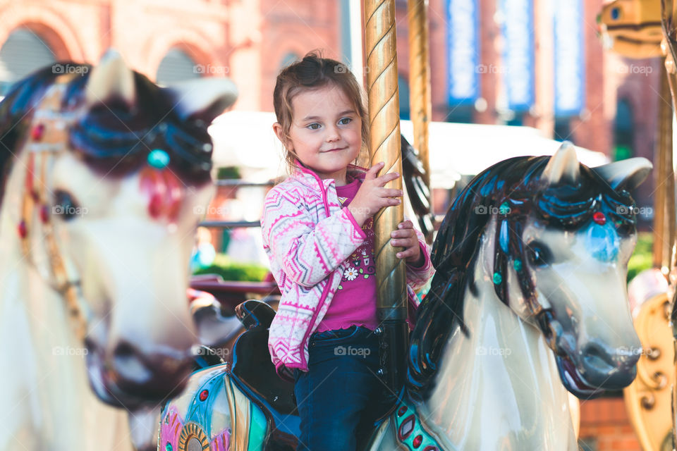 Little adorable smiling girl riding a horse on roundabout carousel at funfair