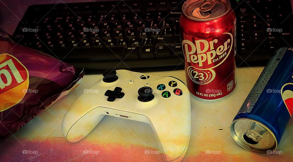 On the table: a black keyboard, a game joystick, a pack of Lays chips, a can of Pepsi, a can of Dr.  Pepper