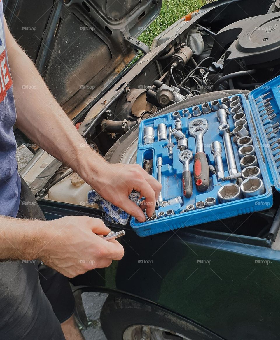A mechanic and his box with metal tools under the hood of an old car
