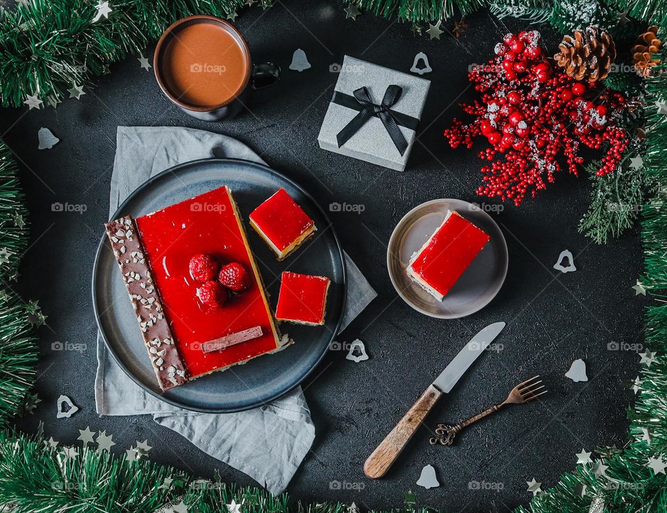 Delicious raspberry cake with berries and slice in saucer with gift box, mug of hot coffee and christmas decoration on dark stone table, flat lay closeup. Holiday food concept.