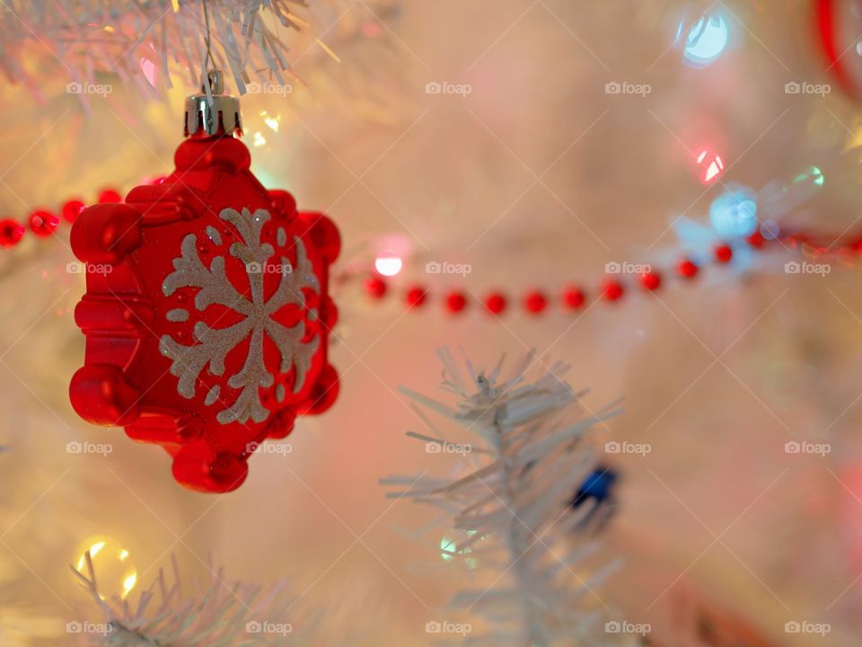 A pretty red star shaped ornament with a white glitter snow flake on it hangs from a white Christmas tree. 