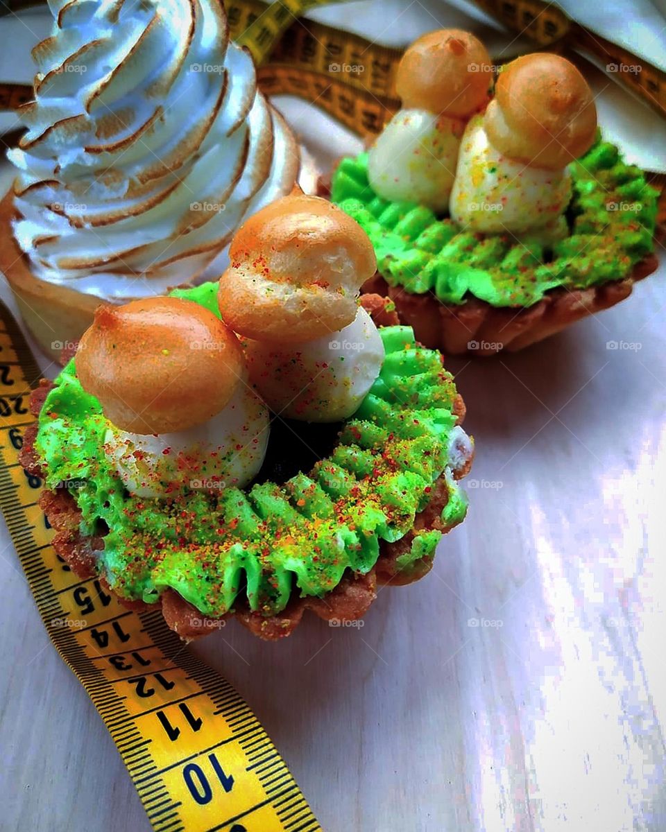 Green color. There are three cakes on a white background. Two cakes with green cream and biscuit mushrooms. The third cake is a basket of bizet cream. Around the cakes lies a yellow centimeter tape