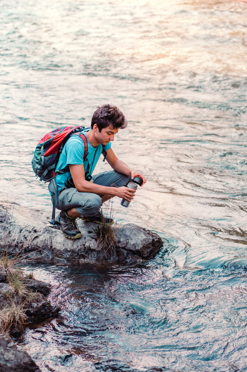 Young wanderer takes pure water from a river into mug. He is sitting on a rock over the river, rests during a hike, spends a vacation on wandering with backpack, he is wearing sport summer clothes