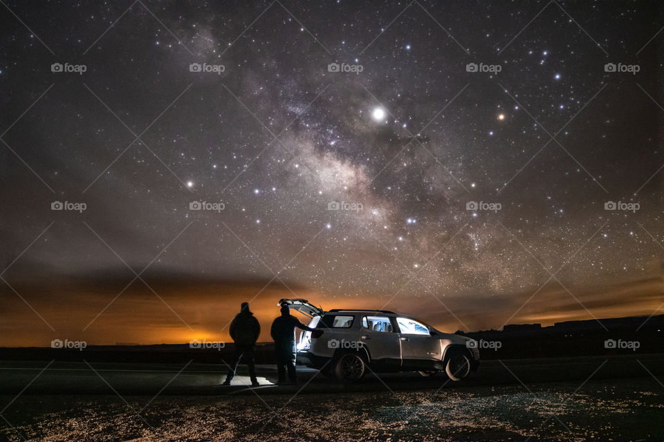 Silhouettes of friends standing by their car gazing up at the Milky Way in the starry night sky. 