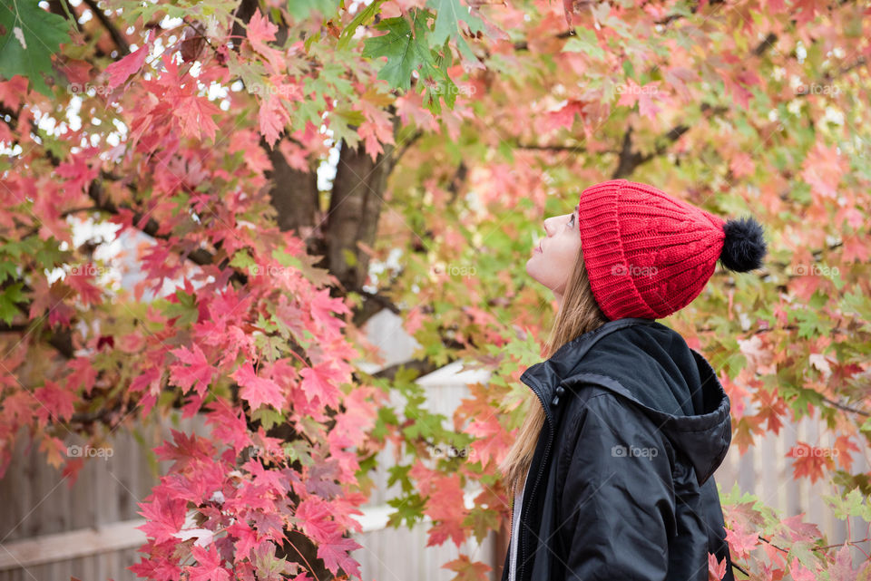 Young millennial woman wearing a red beanie hat and looking up at a tree with fall colored leaves