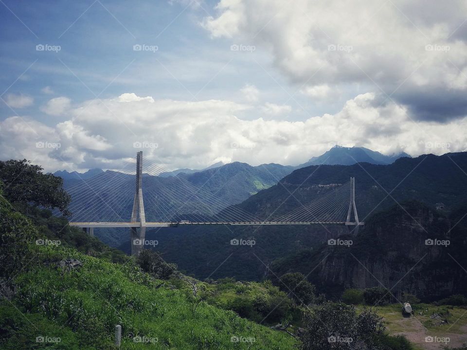 Long bridge crossing between two mountains with more mountains and a cloudy sky as background.