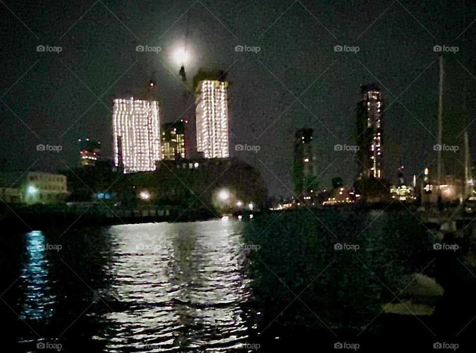 A panoramic shot of two high rise buildings across the “East River” at “Newtown Creek” in LIC, Queens. The night illumination of the two colossal structures w a bright full moon above them is quite stunning. 2022. Hypnotic Productions