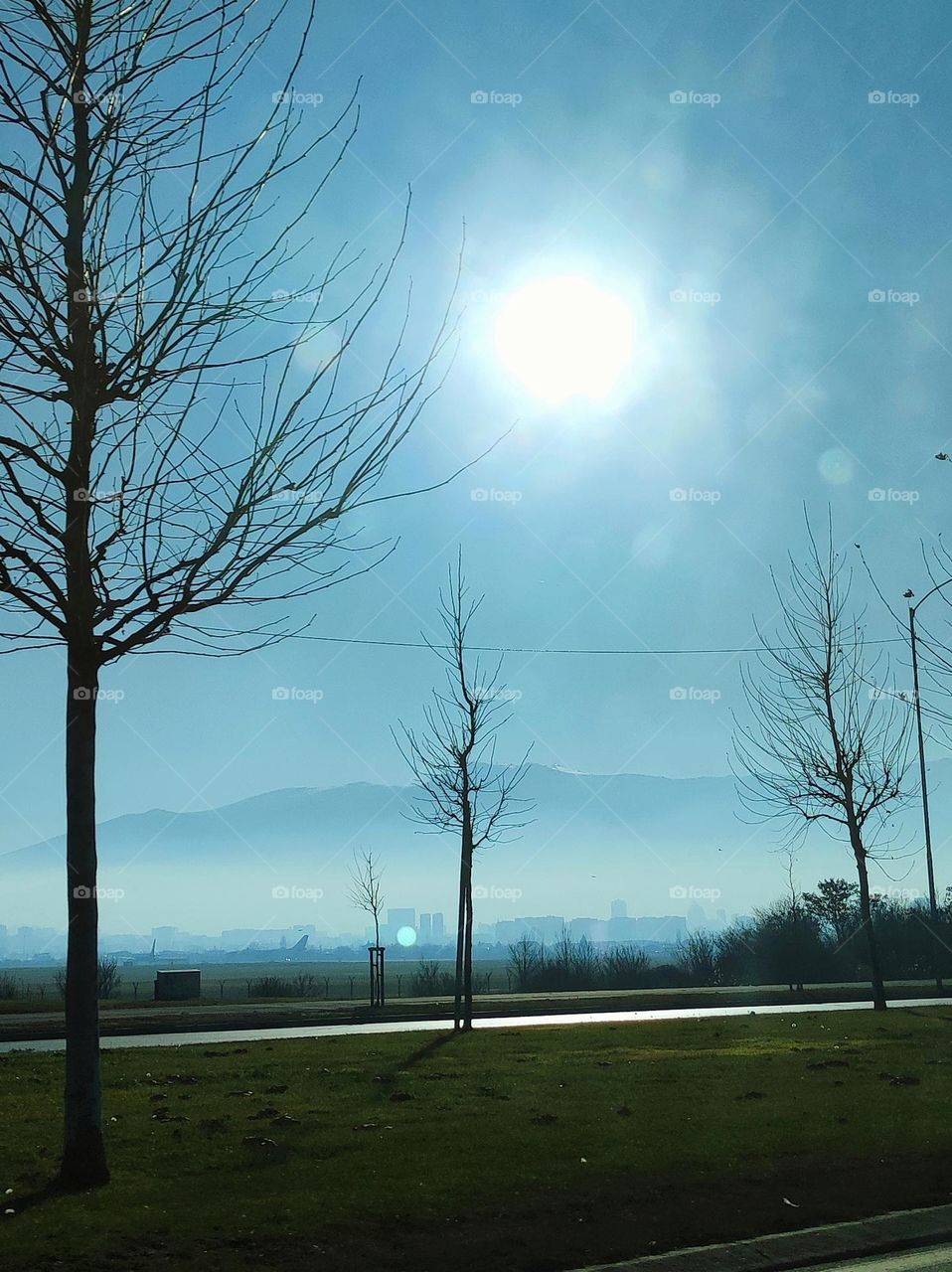 A street with trees and foggy mountains in the back, a shining sun and the buildings of a city and its airport in the fog