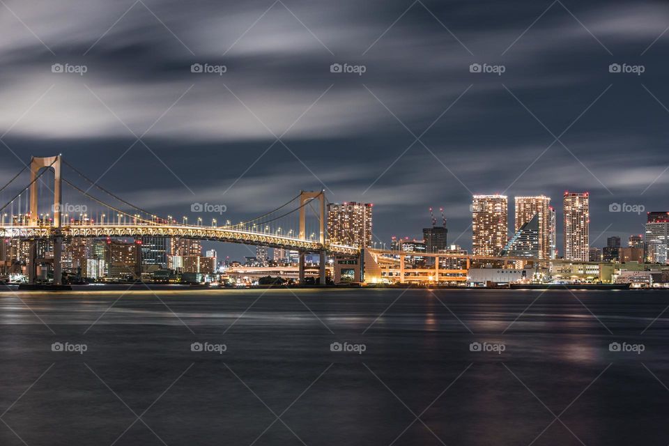Rainbow Bridge over Tokyo Bay at night captured in long exposure. Movements of clouds and water. Office, business, and residential high rise buildings make the skyline of this outer parts of the Japanese capital city.
