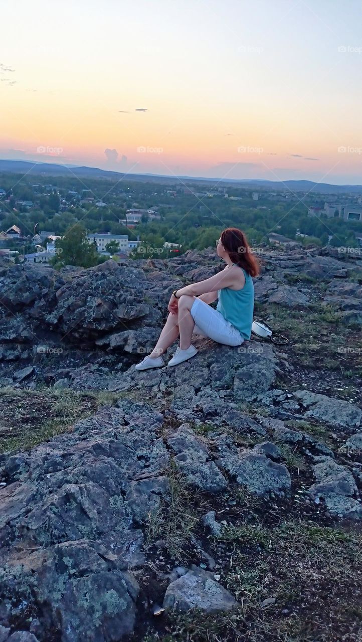 girl looks into the distance, meditation, contemplation nature, view from the mountain to the city, summer, evening, calmness, peace, light breeze, the girl is sitting on the grass