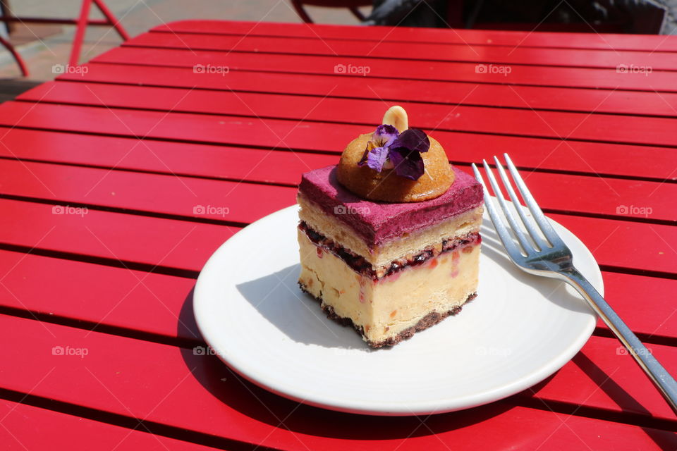 Delicious piece of cake in white plate on top of red wooden table asking to be eaten 