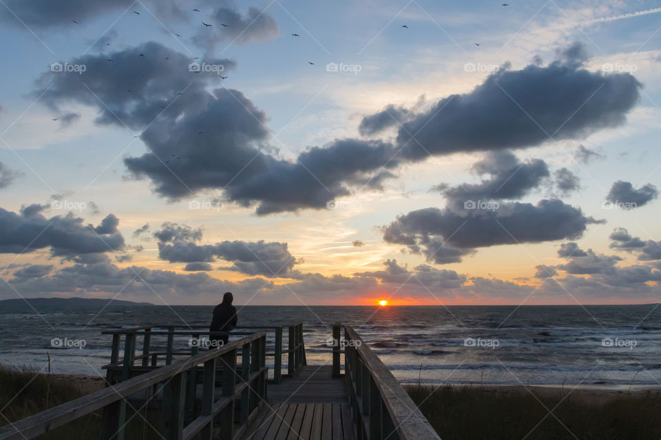 At the end of the day, a man standing on wooden pier watching the sunset by the ocean 