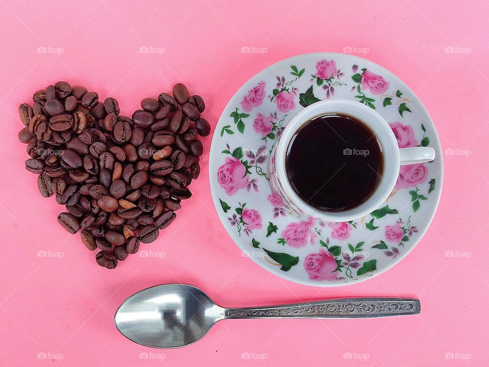 On a pink background, a saucer with pink flowers, a cup of black coffee. Nearby on a pink background lie coffee beans in the form of a heart. Under the heart of coffee beans and a saucer with a cup lies a silver spoon