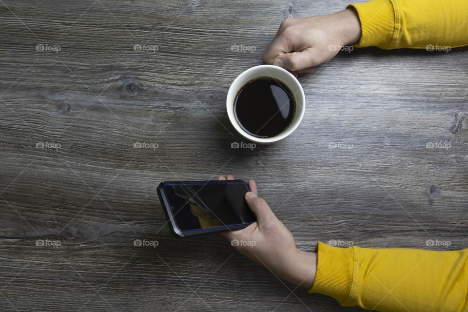 A man drinks coffee in the morning and works on the phone. Men's hands hold a white cup of coffee and a mobile phone in a yellow jacket. The hands lie on a gray wooden surface that is used as a background.