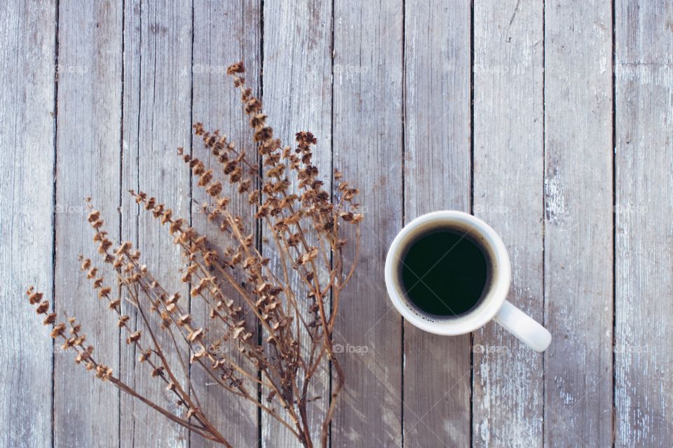 Flat lay of coffee in a white mug next to a dried brown plant on a weathered wooden surface