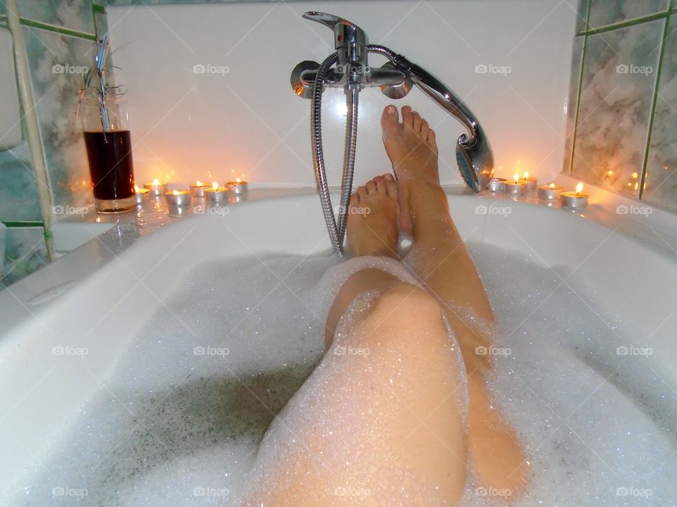 Woman's legs covered in foam while taking a bubble bath with candles and a glass of Coca Cola
