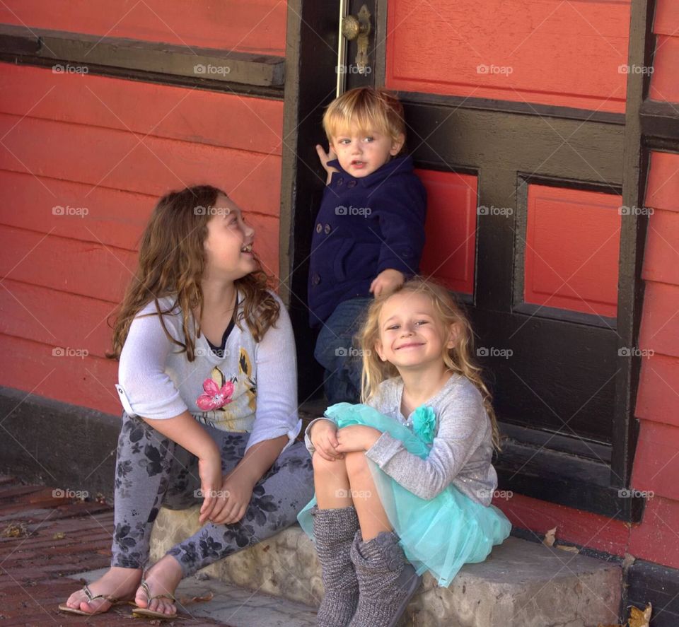 Three adorable siblings gathered outside of a red door at the train station. 