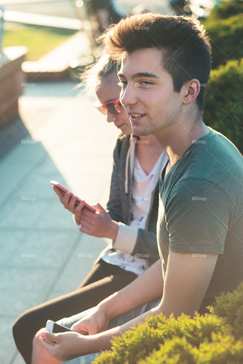 Couple of friends, teenage girl and boy, having fun together with smartphones, sitting in center of town, spending time together. Real people, authentic situations