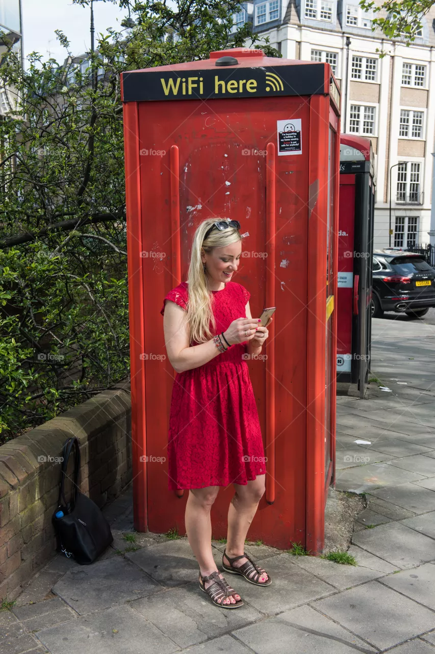 Woman 30 years old looking at her phone and social media at at WiFi spot at a telephone booth in London near Euston Square.