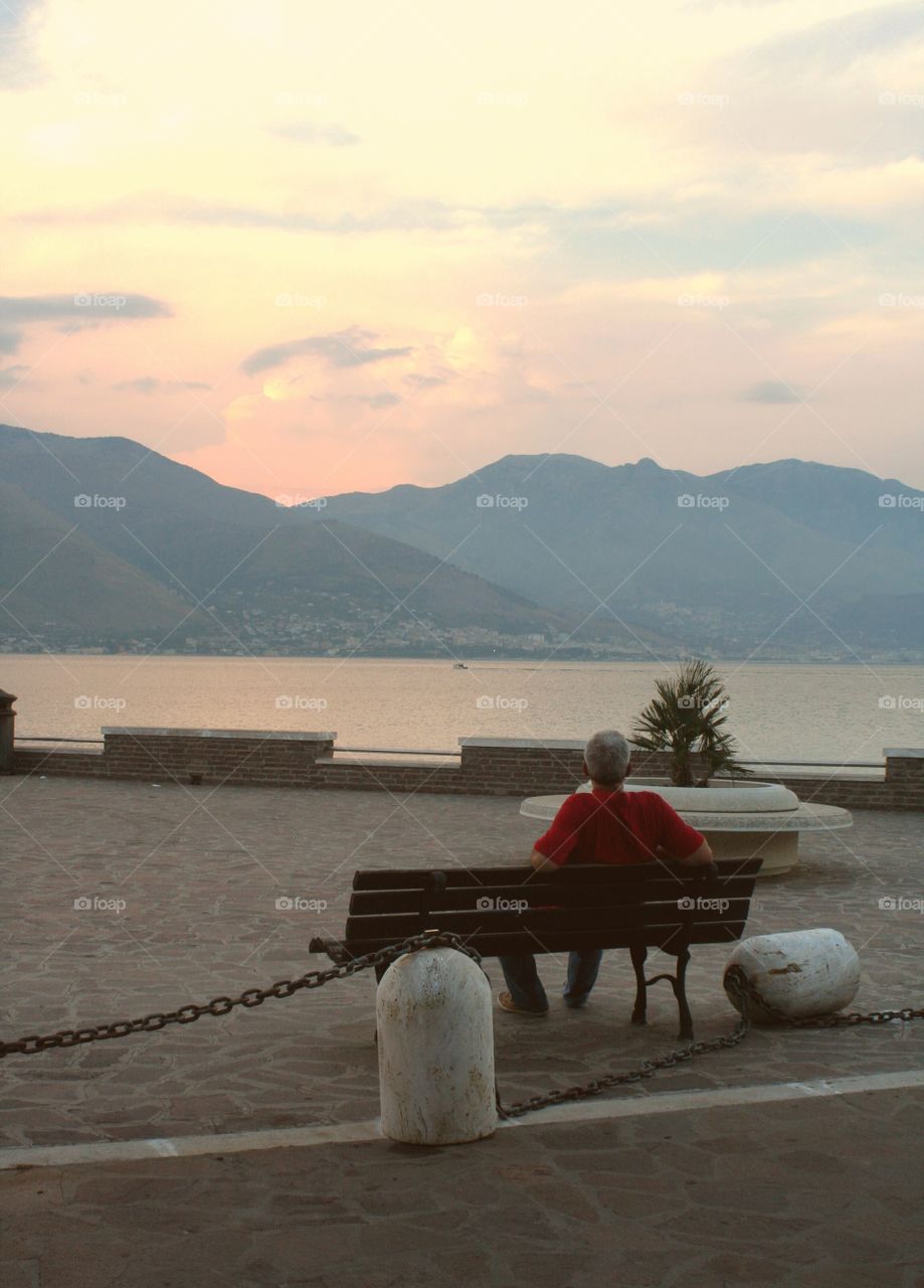 Old man in red tshirt relaxes on bench on seawalk during evening.