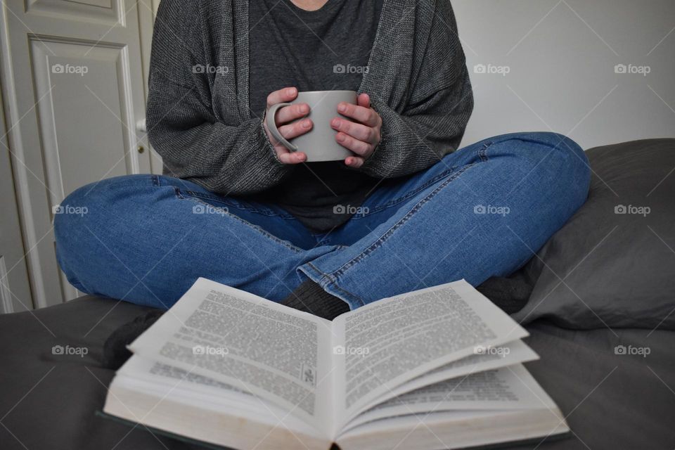 Woman sitting on the bed, while drinking coffee and reading a book. Holding a coffee mug or cup. Woman studying. Wearing denim jeans and grey jersey. White walls, grey and blue clothing. Hand, legs and fingers. Grey T-shirt and comfortable jeans.