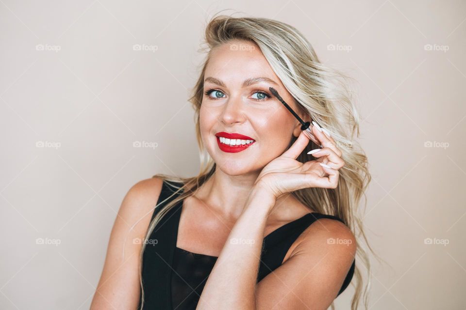 Portrait of gorgeous happy blonde young woman with bright makeup with eyebrow mascara brush in evening dress on beige background
