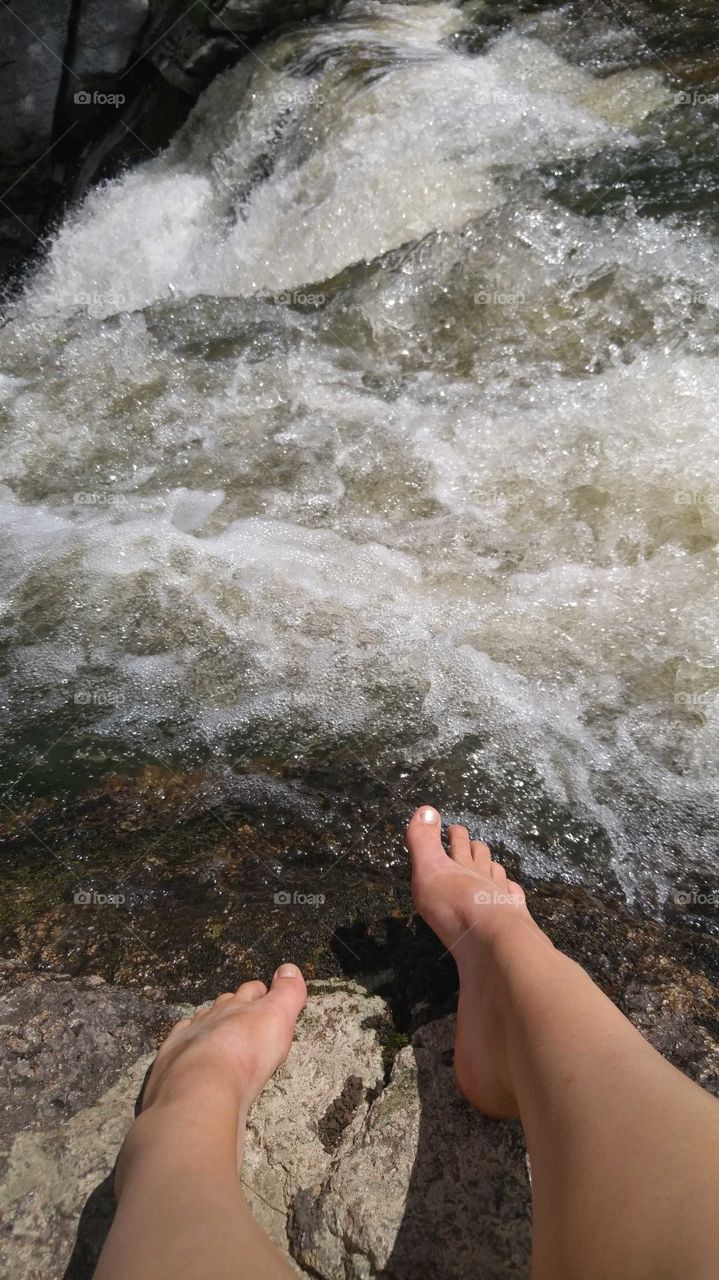 Young woman's feet resting on rough rocks while hanging over the edge of a raging waterfall.