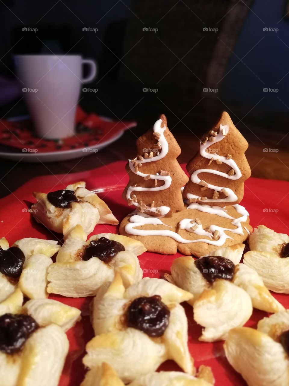 A star shaped red plate full of small pastries with plum jam. A gingerbread decoration with two sugary coated trees with golden pieces. A coffee cup on the multicoloured napkin on a white plate.