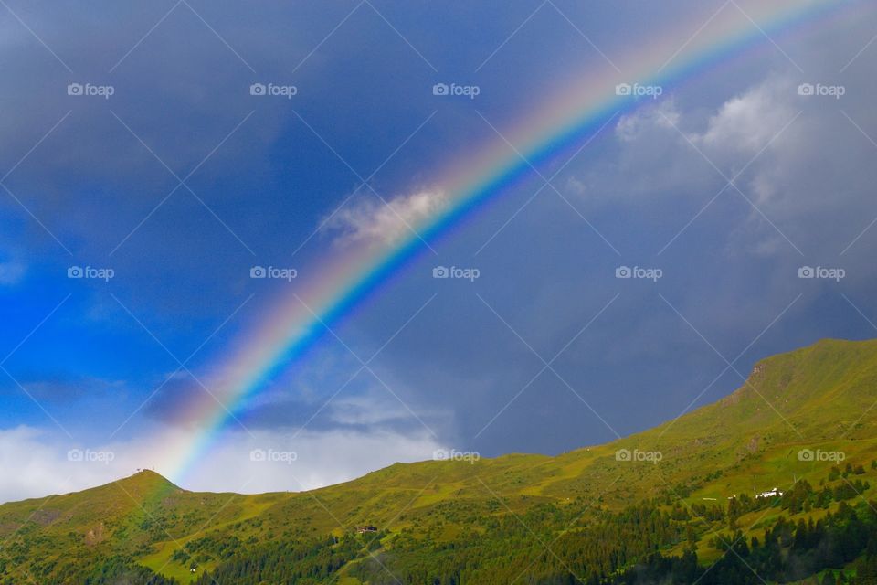 A beautiful rainbow behind a clear green gorgeous hill and in front of a dark sky making the rainbow more clear. 