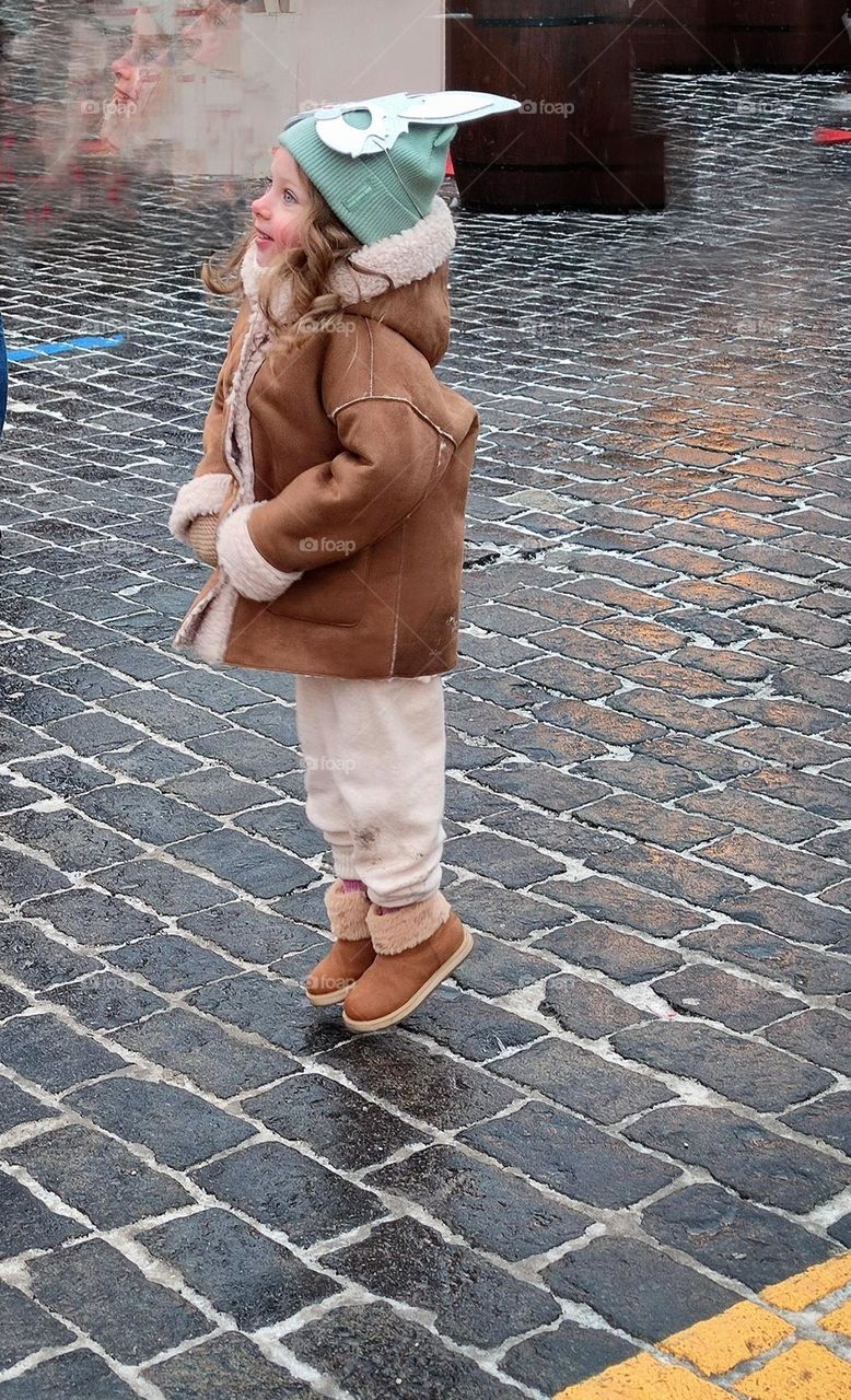 Winter day. On the paving stones of the square, a little girl jumps for joy. the little girl's cap is wearing a paper bunny mask. In the background, a close-up, but blurry, depicts the joyful face of a little girl. Sincere joy of childhood