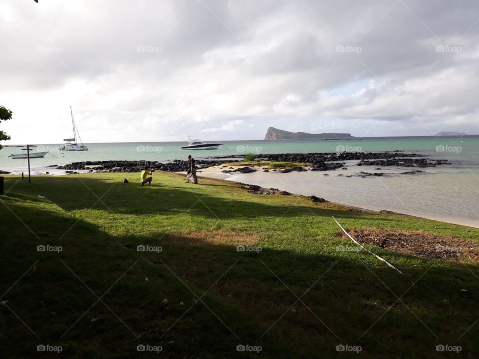 A View at the Seaside with Grass on the ground, Sand at the Front, Green Water, Boats, Island near the Horizon and Tourist taking Photos.