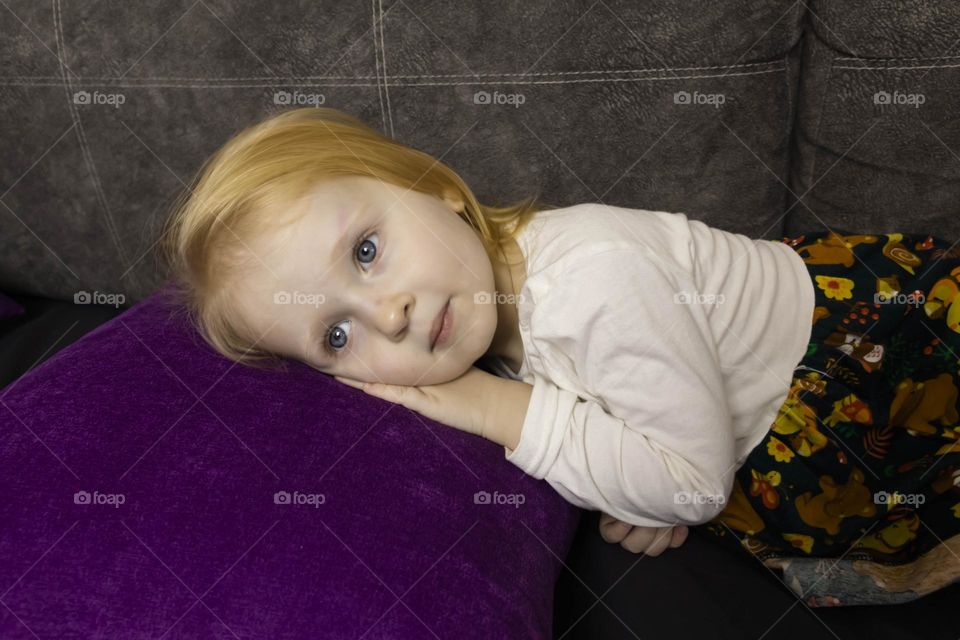 A small, red-haired girl is laughing at home on a gray sofa with purple pillows and a yellow background