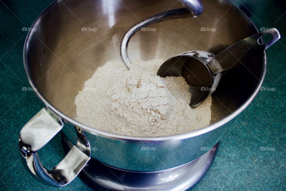Whole-grain four and stainless steel measuring cup in mixing bowl of standing mixer fitted with dough hook on green countertop 