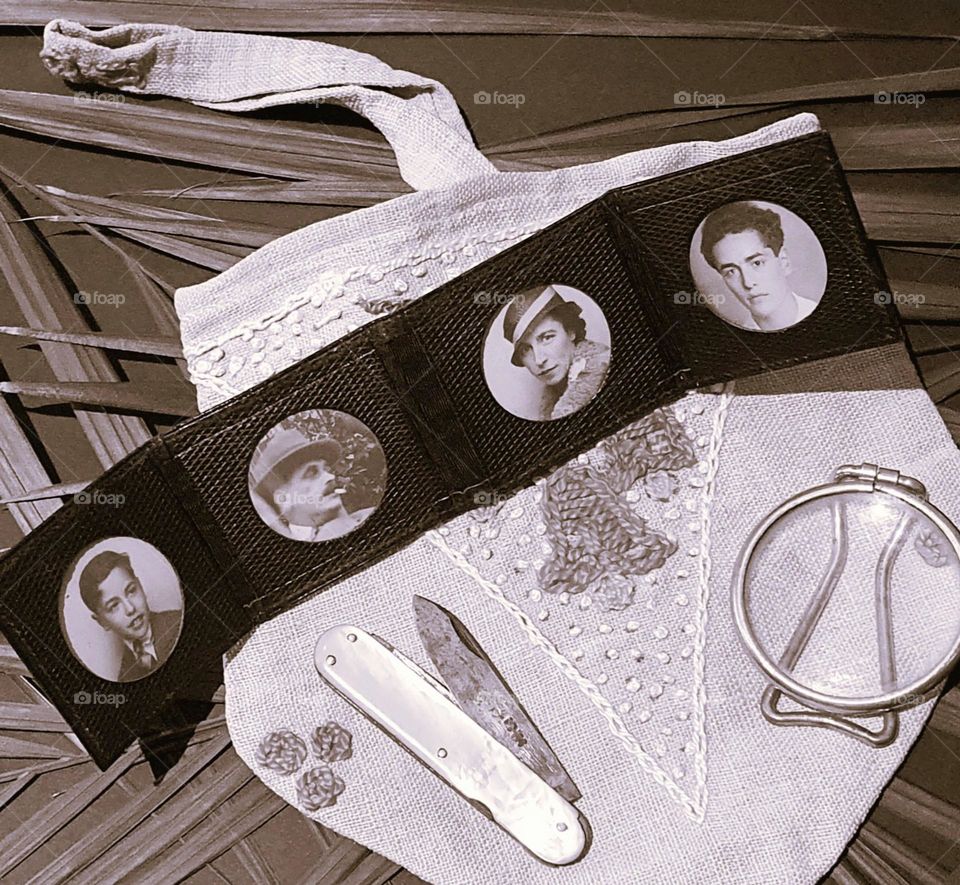 Sepia shot of a vintage flatlay placed on a dark surface and palm leaves composed of a handmade handbag, a mother-of-pearl penknife, a pocket magnifying glass and pocket smug shots photo album with four photos.