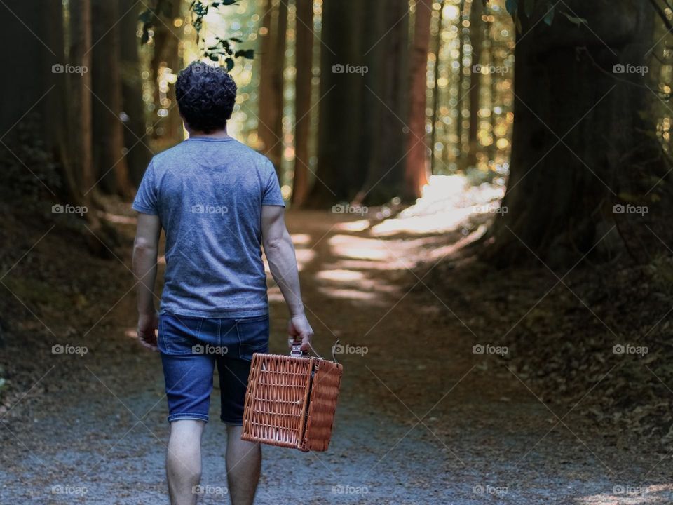 One young caucasian brunette man with curly hair from the back walks along a forest path holding a wicker picnic basket in his hand, close-up side view.