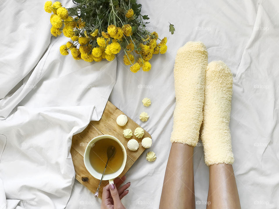 Young woman in cozy yellow socks drinking tea from yellow cup with yellow candies in white cozy bed with yellow flowers 